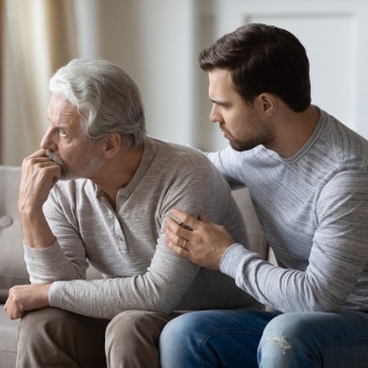 Zwei Herren auf einem Sofa, einer stützt den anderen mit einer Umarmung – Symbolbild für Unterstützung und Fürsorge