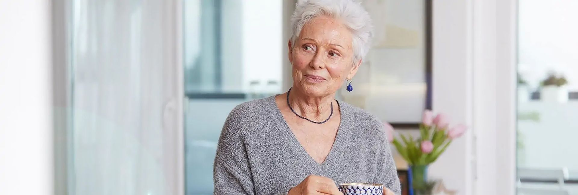 Eine ältere Frau hält eine Tasse mit Untertasse in einem Wohnraum und trägt einen grauen Strickpullover.