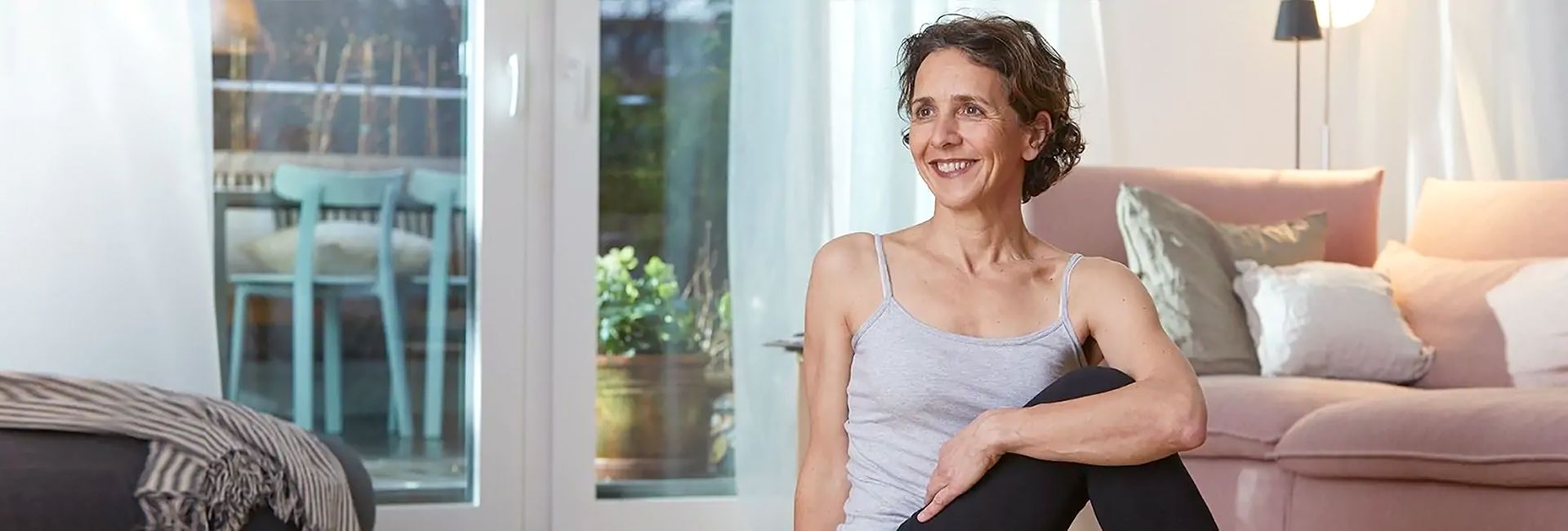 Banner Bild: Eine Frau sitzt entspannt im Wohnzimmer auf dem Boden. Sie hat ein Knie angewinkelt und streckt sich leicht, um ihren Körper zu dehnen. 