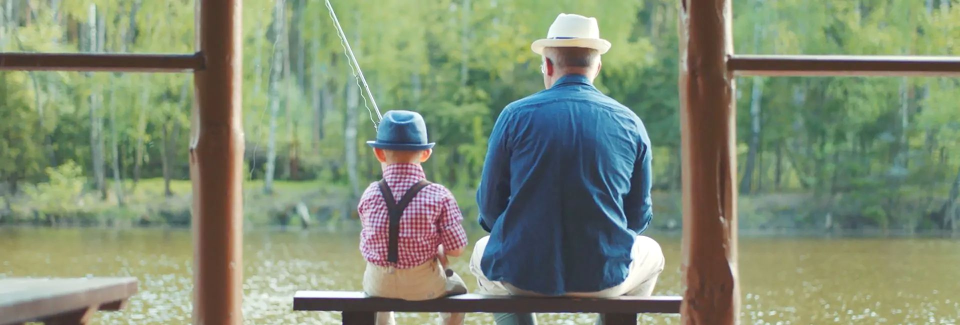 Bannerbild: Ein Mann und ein Kind sitzen mit dem Rücken zur Kamera auf einem Holzsteg an einem See und angeln, umgeben von ruhiger Natur und Bäumen.