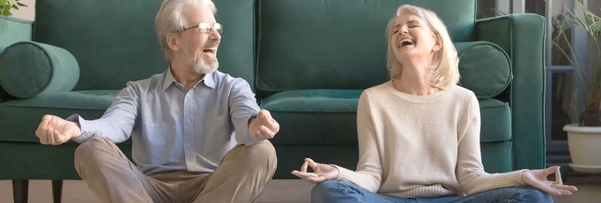 Älterer Herr mit Stoma und Frau sitzen lachend vor einem grünen Sofa im Schneidersitz und meditieren gemeinsam. 
